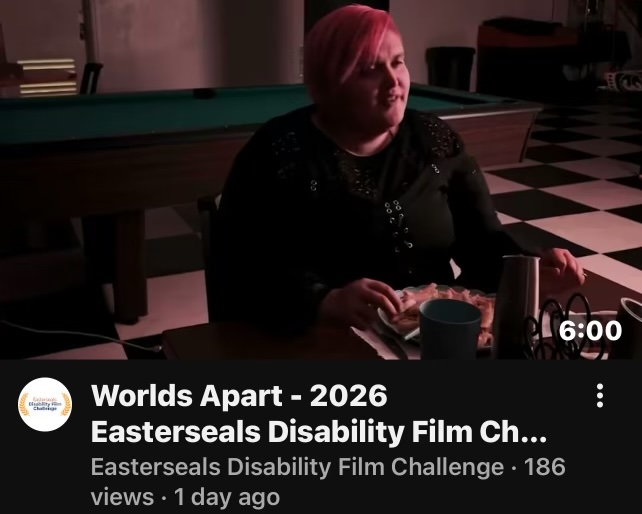 Sloan is sitting in a dimly lit room, at a dining table with plates and silverware in front of them. Sloan is played by Satin LaRance.